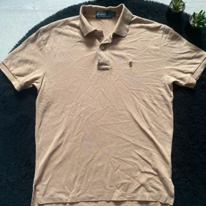 Ralph Lauren pikétröja - Säljer en klassisk beige pikétröja från Polo Ralph Lauren med broderad logga på bröstet. Tröjan har korta ärmar, krage och knäppning med två knappar. Tillverkad i mjuk bomull som är skön mot huden. Perfekt för en clean och stilren look.