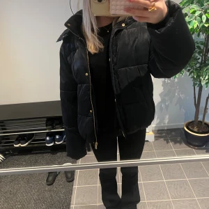 Marinblå pufferjacka med huva - Säljer en Marinblå vinterjacka med huva och guldfärgade detaljer på dragkedja och knappar. Jackan är vadderad, perfekt för kalla dagar. Klassisk look med stora fickor och hög krage. Super varm och skön! Jätte fin i materialet också! Nypris ca 1800kr!