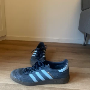 Adidas Spezial mörkblå sneakers - Adidas Spezial sneakers i mörkblå mocka med ljusblå ränder och detaljer. Klassisk rund tå, platt gummisula och snörning framtill. Ikonisk retrostil med guldfärgad Spezial-logga på sidan. Perfekt för dig som gillar streetwear och sportig vibe.