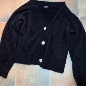 Fluffig ONLY Croppad Kofta Strl M - V-ringning & Diamantknappar - Artikel: ONLFREJA PIUMO ZL L/S CARDIGAN CO KNT 15279599 Säljer en figurnära, fluffig och mjuk kofta från ONLY, storlek M. ​Denna svarta kofta har en trendig kort (cropped) längd och en V-ringning. Det mjuka materialet (fluffigt garn, troligen angora-/mohairimitation) gör den mysig och bekväm. ​Den mest iögonfallande detaljen är de tre stora, glittriga "diamant"-knapparna (faux-rhinestone) som ger koftan en lyxig och festlig touch. 