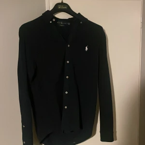 Mörkblå skjorta från Ralph Lauren M - Mörkblå långärmad skjorta från Ralph Lauren i lätt mesh-bomull. Klassisk design med vit broderad logga på bröstet och knappar framtill. Perfekt för dig som gillar stilrena och tidlösa plagg med premiumkänsla.