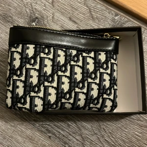  kuvertväska i svart och beige - Snygg kuvertväska med ikoniskt svart och beige mönster, guldfärgad dragkedja och detaljer i svart skinn. Perfekt storlek för det viktigaste och en lyxig touch till din outfit.