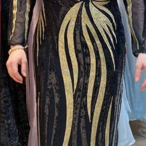 Svart långklänning med guldpaljetter - Svart långklänning med transparenta långärmar och glittrande guldpaljetter i ett coolt, vågigt mönster. Klänningen har en figurnära passform och är tillverkad i polyester med meshdetaljer. Perfekt för dig som vill sticka ut med en glam look.