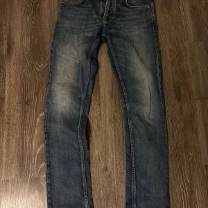 Nudie Jeans Grim Tim, W30 L32 - Säljer ett par klassiska blå jeans från Nudie Jeans med snyggt slitna detaljer och orange sömmar. Modellen har raka ben, fem fickor och knappgylf. Jeansen är tillverkade i bomull och har en cool tvättad look med Nudie-logga bak i midjan.