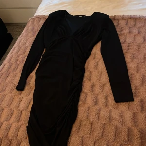 Svart långärmad klänning Bik Bok - Snygg svart långklänning från Bik Bok med långa ärmar och draperad detalj längs sidan. Klänningen har en figurnära passform och en stilren, enkel design som ger en elegant look. Perfekt för dig som gillar klassiska och tidlösa plagg.