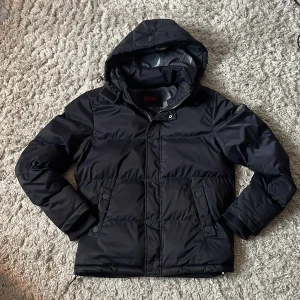 Svart pufferjacka från Guess med huva - Svart pufferjacka från Guess med huva och dragkedja. Jackan är vadderad med syntetisk ecodown, har två fickor med knappar och är vindtät samt vattenavvisande. Perfekt för kalla dagar och har en stilren look med diskret logga på ärmen.