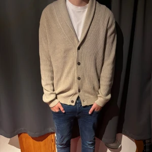 Beige ribbad kofta med knappar från John Henric  - Säljer en beige ribbad kofta från John Henric med stora knappar och bred krage. Koftan har lång ärm och en avslappnad passform, perfekt att slänga på sig över en t-shirt. Enkel och stilren design som funkar till det mesta.