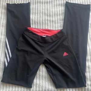 Svarta bootcut Adidas leggings med vita ränder - Svarta leggings från Adidas med klassiska vita ränder längs benen och rosa logga framtill. Insidan av midjan är fodrad i rosa. Tillverkade i ett stretchigt syntetmaterial som passar perfekt för träning eller chill. Snygg och sportig look med tight passform.