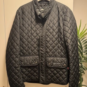 Belstaff Jacka - Belstaff Wilson Lightweight Quilt – Svart Ny skick, knappt använd. Inköpt för 4499 kr. 	•	Storlek: 50 (passar M/L) 	•	Klassisk lätt quiltad jacka i tidlös design 	•	Två fickor med knappstängning + innerficka 	•	Diskret logga framtill, Union Jack-detalj i sidan 	•	Perfekt som vår/höstjacka  Pris: 1299 (nypris 4499 kr)