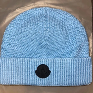 Ljusblå stickad mössa från Moncler - Snygg ljusblå stickad mössa från Moncler med klassisk ribbad uppvikning och svart logomärke framtill. Tillverkad i mjuk bomull som håller dig varm och ger en clean look. Perfekt accessoar för kyliga dagar och enkel att matcha med olika outfits.