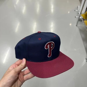 Philadelphia Phillies snapback keps - Snygg snapback keps med Philadelphia Phillies-logga i rött och vitt framtill. Marinblå krona, vinröd skärm och grönt under skärmen. Justerbar baktill för perfekt passform. Perfekt för dig som gillar sportig streetstyle.