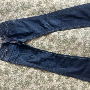 Blå jeans från Nudie Jeans med rå kant - Säljer ett par klassiska blå jeans från Nudie Jeans med snygga kontrastsömmar och rå, fransad kant nertill. Jeansen har fem fickor och en rak passform. Midjemått (mätt rakt över) 32cm, Innerbenslängd 74cm