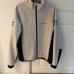  vindjacka från Sail Racing - Snygg vit vindjacka från Sail Racing med svarta detaljer längs sidorna och ärmsluten. Jackan har hög krage, hel dragkedja framtill och logga på bröstet samt ärmen. Perfekt för dig som gillar stilren och sportig look.
