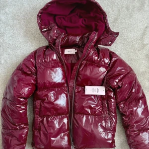 Vinröd pufferjacka från Nelly - Snygg vinröd pufferjacka från Nelly med glansig finish och huva. Jackan har dragkedja framtill, stora fickor och quiltad design. Perfekt för kalla dagar och ger en cool vibe till din outfit.