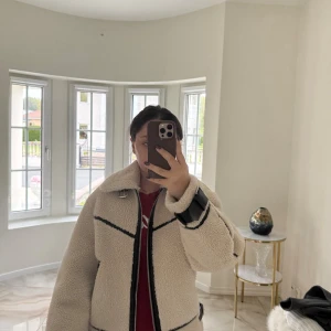 Beige teddyjacka med svarta detaljer - Säljer en beige teddyjacka med svarta detaljer och dragkedja framtill. Jackan har stora slag, breda ärmar och coola spännen vid handlederna. Perfekt för dig som vill ha en mysig och trendig look under höst och vinter.