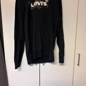 Svart hoodie från Levi's med camo-logga - Svart hoodie från Levi's i storlek L med klassisk kamouflagemönstrad logga på bröstet. Tröjan har huva, magficka och långa ärmar. Tillverkad i mjuk bomull, perfekt för chill dagar. Snygg detalj med röd Levi's-tag på sidan.