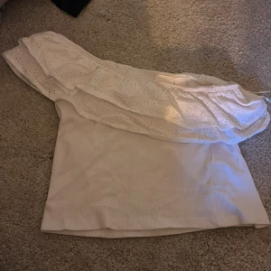 Vit offshoulder topp med volang Gina Tricot - Supersöt vit offshoulder topp från Gina Tricot med bred volang upptill och broderade hålmönster. Toppen är ärmlös och har en ribbad struktur i tyget. Perfekt för dig som gillar romantisk och feminin stil.