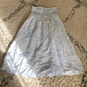 Ljusblå långkjol med snörning - Säljer en ljusblå långkjol med bred smock i midjan och dekorativ snörning med tofsar framtill. Kjolen har en luftig och flowy passform, perfekt för varma dagar. Materialet känns lätt och bekvämt mot huden.