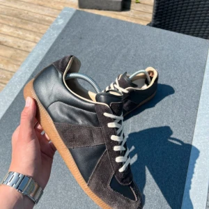 Maison Margiela sneakers svart mocka - Maison Margiela sneakers i svart skinn och mocka med beige detaljer och klassisk gummisula. Snygg retrostil med vita snören och rund tå. Perfekta för dig som gillar stilrena och tidlösa sneakers med lyxig känsla. Skriv om du undrar något !