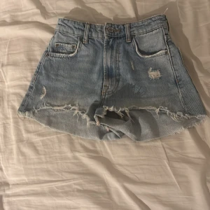Ljusa jeansshorts från Zara - Säljer ett par ljusblå jeansshorts från Zara med råa, fransiga kanter och slitna detaljer. Klassisk femficksmodell med en högre midja så helt perfekta att vika ner. Pris kan ev. diskuteras 