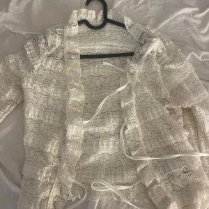 Vit spetsig bolerotopp med volanger - Säljer en vit bolerotopp i spets med långa ärmar och snygga volangdetaljer. Toppen har knytband framtill och är helt genombruten med ett romantiskt mönster. Perfekt för dig som gillar en feminin och trendig look.