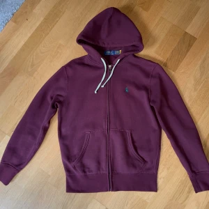 Vinröd hoodie från Polo Ralph Lauren - Vinröd hoodie från Polo Ralph Lauren med dragkedja, huva och vita snören. Klassisk broderad logga på bröstet och två fickor framtill. Perfekt för chill dagar och snygg till jeans eller joggers. Xs