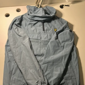Blå vindjacka från Lyle & Scott - Snygg ljusblå vindjacka från Lyle & Scott med klassisk gul logga på bröstet. Jackan har huva, halv dragkedja och meshfoder på insidan. Perfekt för blåsiga dagar och enkel att matcha med streetwear.