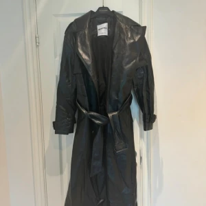 Svart trenchcoat i skinn från Bershka - Säljer en svart trenchcoat i skinnimitation från Bershka. Jackan har bälte i midjan, stora slag och spännen vid ärmsluten. Lång modell med klassisk trenchcoat-look, perfekt för dig som vill sticka ut med en edgy vibe.