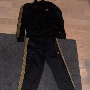 Svarta EA7 mjukisbyxor med guldiga ränder - Säljer ett par svarta mjukisbyxor från EA7 Emporio Armani med guldiga ränder längs sidorna och logga på benet. Byxorna har snörning i midjan och är gjorda i ett glansigt syntetmaterial som känns riktigt sportigt och snyggt.