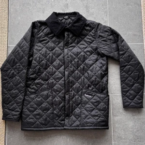 Svart quiltad jacka från Barbour - Svart quiltad jacka från Barbour med klassisk krage i manchester. Jackan har tryckknappar framtill, två stora fickor och ett stilrent rutmönster. Perfekt för dig som vill ha en clean och tidlös look under höst och vår. Jackan är i strl XL (barn) vilket motsvarar ca 14-16år beroende på kroppsbyggnad. 