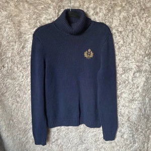 Mörkblå stickad polotröja från Ralph Lauren - Mörkblå stickad polotröja från Ralph Lauren med broderad guldfärgad logga på bröstet. Tröjan har ribbade muddar och hög, tjock polokrage. Tillverkad i 100% bomull och har en klassisk, tidlös look.