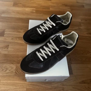 Svart/bruna   Maison Margiela gata  - Snygga svarta sneakers från Maison Margiela med vita kontrastsömmar och vita snören. Skorna har en klassisk låg siluett, rund tå och beige sula. Tillverkade i skinn och mocka för en lyxig känsla. Perfekta för dig som gillar stilrena och tidlösa sneakers.