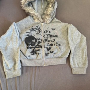 Grå croppad hoodie med tryck från SHEIN - Grå croppad hoodie från SHEIN med svart tryck framtill och texten 'Make Some Crazy Noise'. Hoodien har dragkedja, långa ärmar, mjuk fuskpäls i luvan och bred mudd i midjan. Perfekt för dig som gillar streetstyle och vill sticka ut.