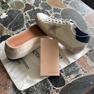 Golden Goose Superstar sneakers vit/blå - Golden Goose Superstar sneakers i vitt skinn med grå mockadetaljer och blå hälkappa. Klassisk stjärndesign på sidan, snörning framtill och platt sula med fiskbensmönster. Snygg kontrast mellan material och färger, perfekt för en trendig streetstyle.