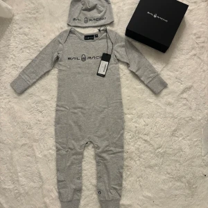 Sail Racing babyset i presentbox 12-18 - Sail Racing babyset, långärmad jumpsuit och mössa i grått. Setet består av en mjuk och bekväm helkroppsdräkt med tryckknappar längs benen samt en matchande mössa med loggan. Storlek 12-18 månader 100% bomull 🌟HELT NY med tag kvar! 🌟 Kommer i original presentförpackning. Perfekt som present! 🩶 Finns i fler färger och storlekar i mina annonser.