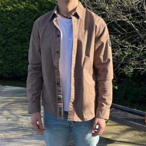 Beige overshirt från Calvin Klein - Säljer en snygg brun/beige overshirt från Calvin Klein Jeans med klassisk krage och svarta knappar. Skjortan har lång ärm och en tydlig CK-logga på vänster ärm. Perfekt att bära öppen över en t-shirt för en chill look.