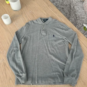 Grå långärmad pikétröja Ralph Lauren - Säljer en stilren grå långärmad pikétröja från Polo Ralph Lauren. Tröjan har klassisk krage, två knappar och den ikoniska broderade loggan på bröstet. Perfekt för en clean och avslappnad look.