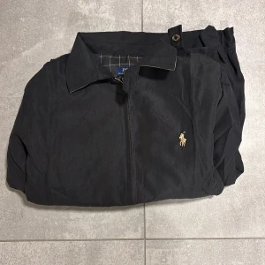 Svart vindjacka från Polo Ralph Lauren - Svart vindjacka från Polo Ralph Lauren med klassisk krage, dragkedja framtill och broderad gul logga på bröstet. Jackan har elastiska muddar i midja och ärmslut samt rutigt innerfoder. Perfekt för dig som gillar stilren och tidlös design. Passar mer som S än M enligt mig.