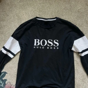 Svart långärmad tröja Hugo Boss M - Svart långärmad tröja från Hugo Boss med vit logga framtill. Ärmarna har vita partier och svarta ränder för en sportig look. Tillverkad i mjuk bomull och har en klassisk passform. Perfekt för dig som gillar stilrena och trendiga plagg.