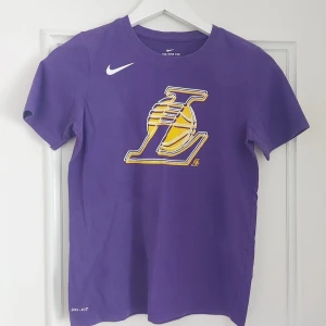 Lila Lakers t-shirt från Nike 13-14år - Snygg lila t-shirt från Nike med stort Los Angeles Lakers-tryck på bröstet och vit swoosh-logga. Tillverkad i Dri-FIT-material som andas och håller dig fräsch. Klassisk passform med rund hals och korta ärmar. Perfekt för basketfans!