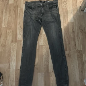 Replay Anbass grå slim jeans - Snygga grå jeans från Replay, modell Anbass med slim fit. Jeansen har klassisk femficksdesign, svarta detaljer och diskret slitning för en cool look. Tillverkade i mjukt jeanstyg som sitter skönt och följer kroppen. Perfekta för dig som gillar stilrena och moderna jeans.