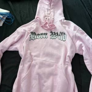 Born Wild rosa hoodie med Betty Boop - Rosa hoodie från Collusion i storlek S med coolt Born Wild-tryck fram och stor Betty Boop-grafik på ryggen. Tröjan har huva med snörning och känguruficka. Mjuk bomullsmix, perfekt för dig som gillar statement-plagg och streetstyle.