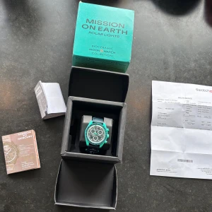 Omega x Swatch mission on Earth klocka  - Säljer en Omega x Swatch Speedmaster Mission on Earth Polar Lights från Bioceramic MoonSwatch Collection. Klockan har en rund boett i turkosgrön färg, svart urtavla med tre subdials och vita detaljer. Bandet är svart med Swatch-logga. Kommer med originalbox och manual.