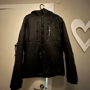 Svart pufferjacka från Sail Racing - Svart pufferjacka från Sail Racing med huva och flera dragkedjeförsedda fickor framtill. Jackan har en sportig look, diskret logga på bröstet och är tillverkad i syntetmaterial. Perfekt för kyliga dagar och enkel att matcha med det mesta.