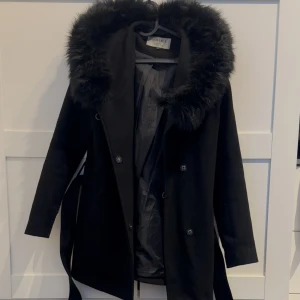 Svart kappa med fuskpäls från Hollies - Svart kappa från Hollies med stor huva kantad av fuskpäls. Jackan har knappar framtill, bälte i midjan och en klassisk siluett. Perfekt för kalla dagar med sin varma känsla och snygga look. Fodrad insida och diskret logga på ärmen.