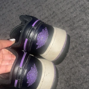 Nike Air Jordan 1 Low svart/lila - Säljer ett par Nike Air Jordan 1 Low i svart och lila med vita sulor. Skorna har klassisk Jordan-logga på hälen och svart snörning. Ovandelen är i skinn och textil, med perforerad tåbox för extra andningsförmåga. Perfekta för dig som gillar streetwear och sneakers med attityd.