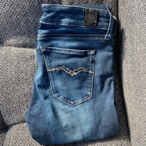 Blå Replay jeans W27 med snygga detaljer - Säljer ett par blå jeans från Replay i storlek W27. Jeansen har klassisk femficksdesign, snygga slitningar och kontrastsömmar på bakfickan. Märkespatch bak i midjan och Replay-logga på fickan. Perfekta för dig som gillar stilrena och trendiga jeans.