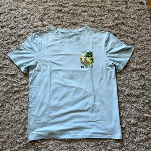 Casa Blanca T-shirt - Snygg ljusblå t-shirt från Casa Blanca med tryck av palmer och byggnader både fram och bak. Klassisk rund hals och korta ärmar. Skön och mjuk känsla, perfekt för sommardagar. Trycket ger en chill vibe och sticker ut på ett stilrent sätt.