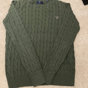 Olivgrön kabelstickad tröja från Gant - Säljer en olivgrön kabelstickad tröja från Gant i 100% bomull. Tröjan har rund halsringning, ribbade muddar och Gant-logga broderad på bröstet. Perfekt för dig som gillar klassisk och stilren look.