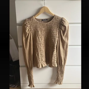 Beige smockad blus med puffärm - Supersöt beige blus från Gina Tricot med smockat tyg och långa puffärmar. Blusen har rund halsringning och volangdetaljer vid ärmslut och nederkant. Perfekt för dig som gillar trendiga och bekväma plagg med lite extra detaljer.